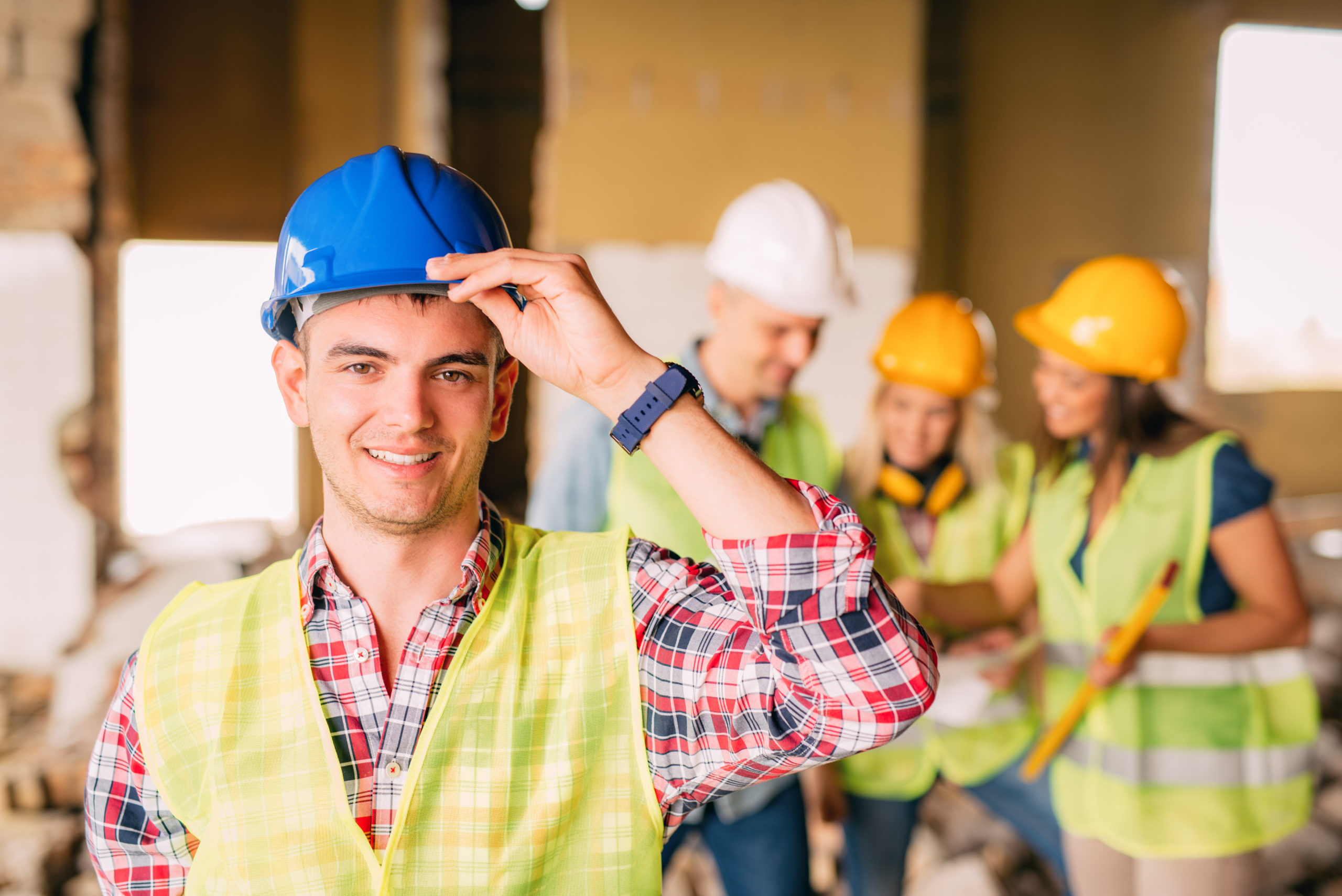 Successful Young Electrician Smiling young electrician holding helmet and looking at camera. His architect colleagues review plan in background at a construction site.